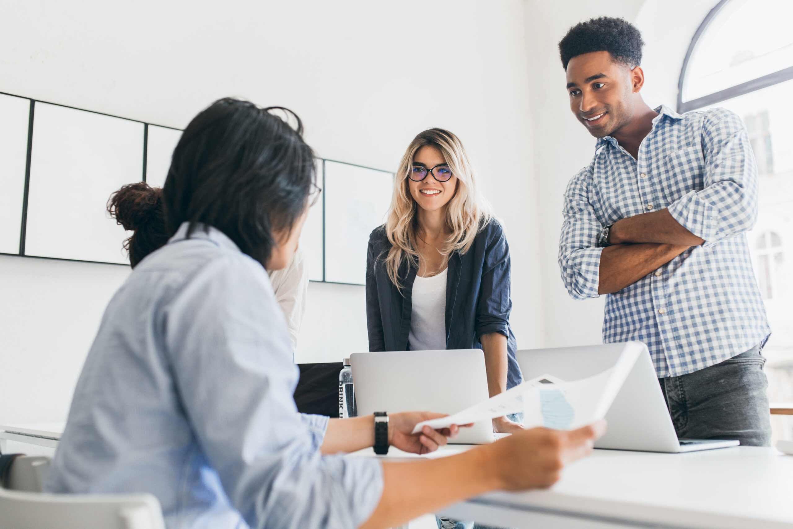 african office worker in checkered shirt standing with arms crossed and looking at asian manager. indoor portrait of freelance web developers discussing something and using laptops..